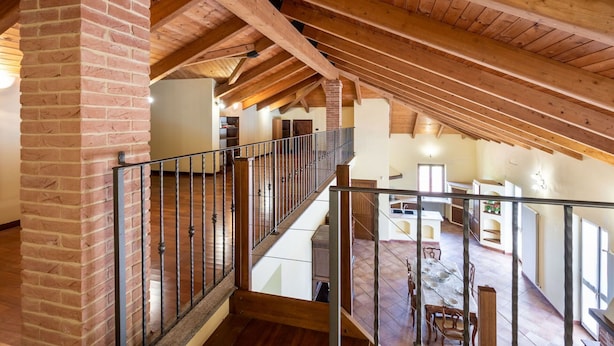 A spacious loft with exposed wooden beams, a brick column, and a balcony overlooking a dining area.