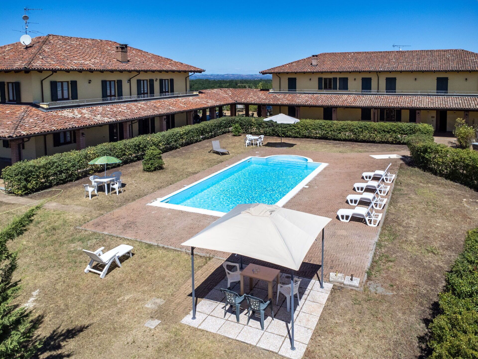 A swimming pool surrounded by lounge chairs and umbrellas, with a covered seating area and a view of two buildings in the background.