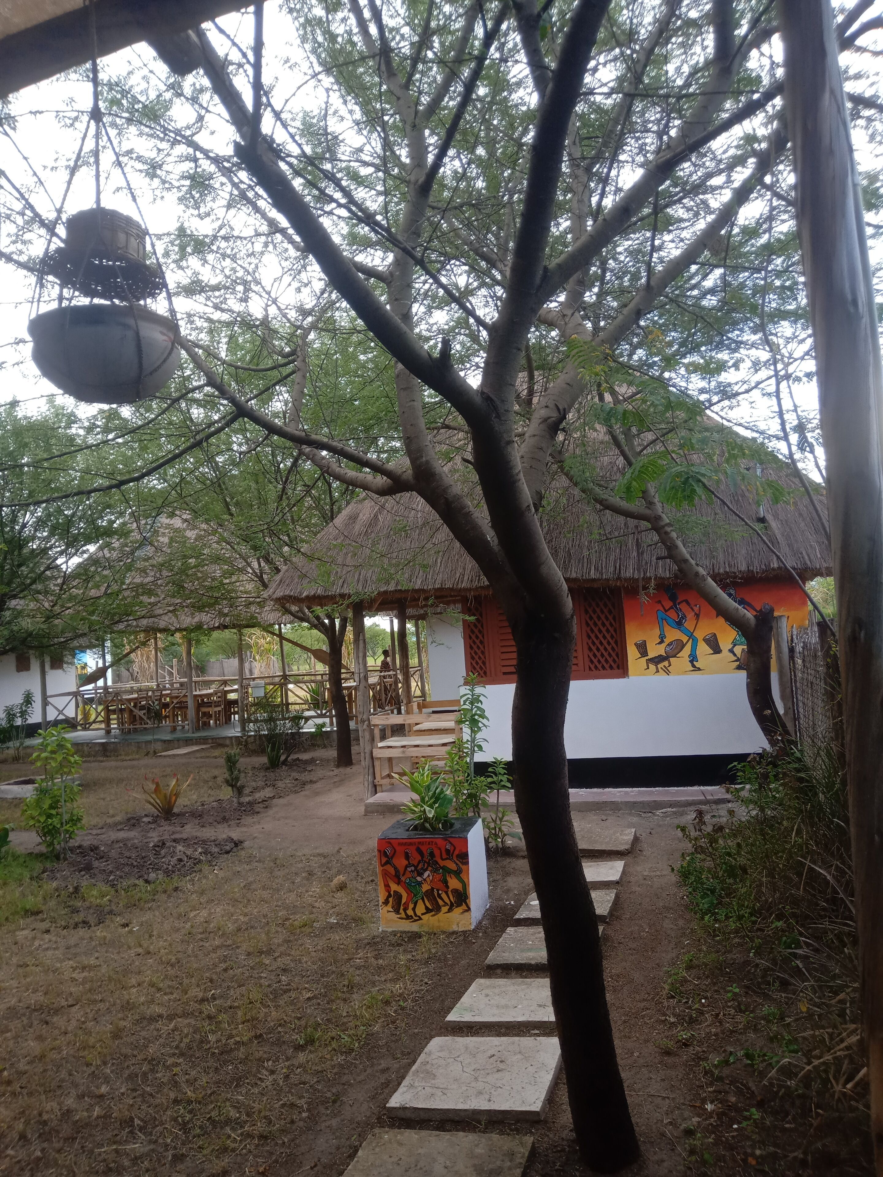 A rustic outdoor area with a thatched-roof structure, a wooden bench, and a pathway leading to a building with a colorful mural.