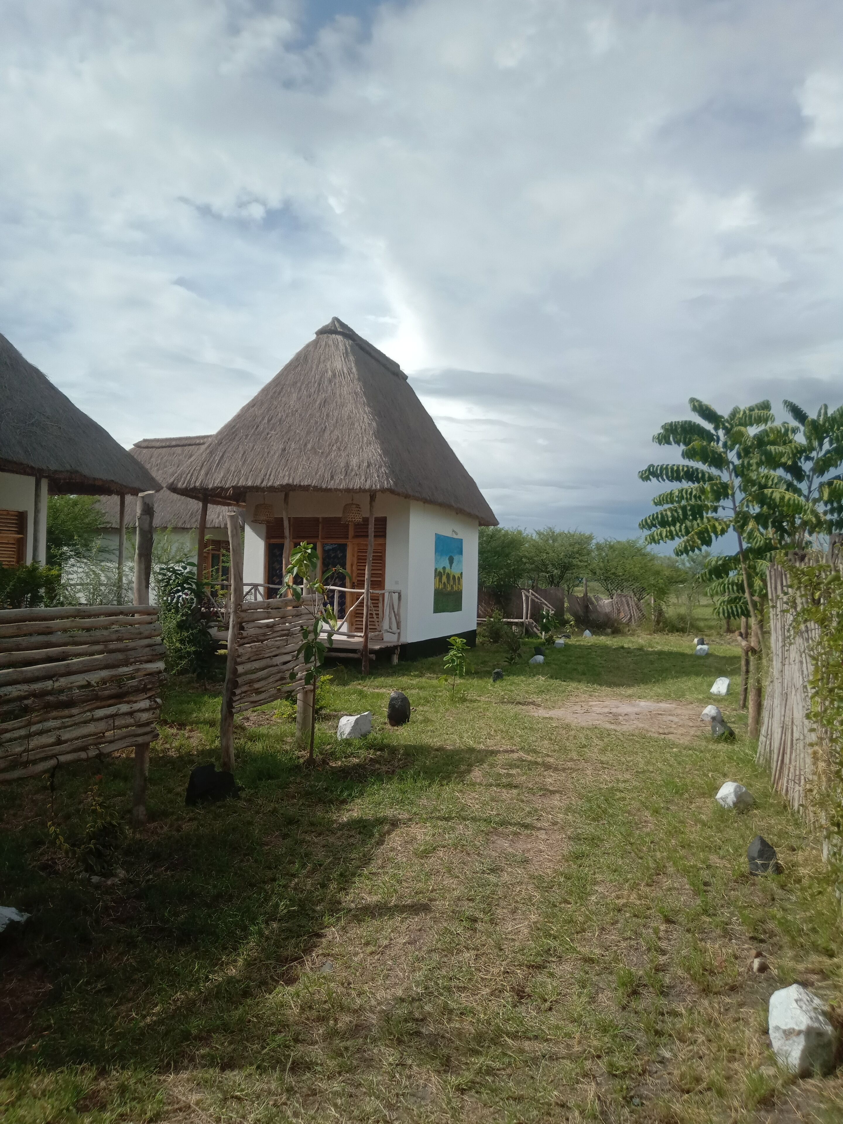 Un bungalow con tetto di paglia e un balcone, circondato da un'area erbosa con rocce sparse e una recinzione in legno.