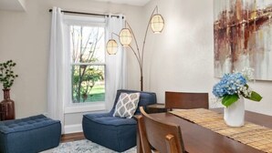 Un coin repas avec une table en bois, des chaises recouvertes d’un tissu bleu, un pouf bleu et un vase avec des fleurs bleues.
