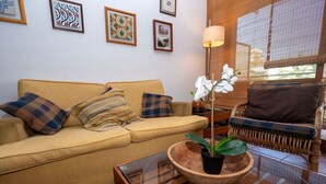 A living room with a yellow sofa, a wooden coffee table, and a wicker chair. There are framed artworks on the wall and a floor lamp.