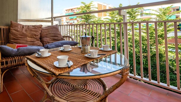 A balcony with wicker furniture, a glass table, and a view of buildings and greenery.
