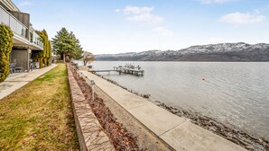 A lakeside property with a dock extending into the water, a grassy area with a stone border, and a building with a balcony overlooking the lake.