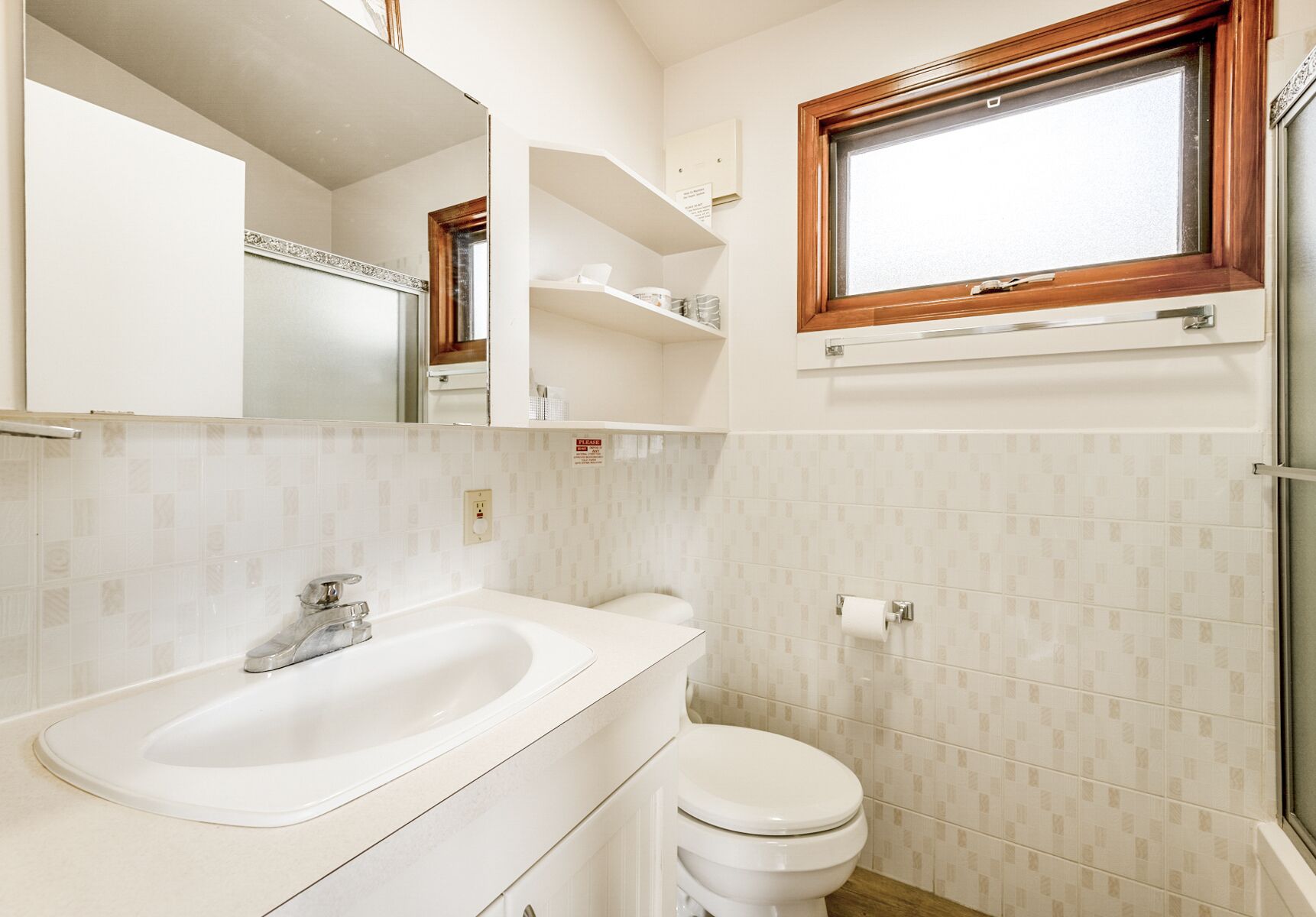 Un baño con lavamanos blanco, espejo y retrete. Se aprecia una ventana con marco de madera sobre el inodoro.