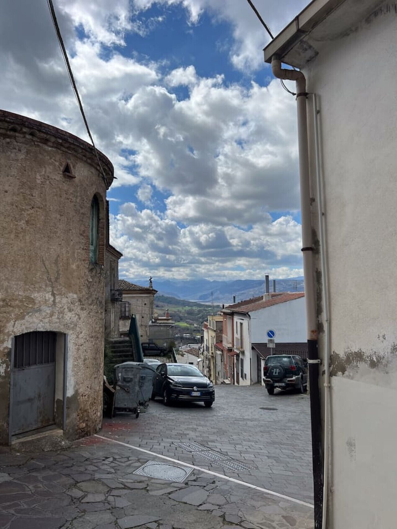 A narrow street with buildings on either side, a parked car, and a clear sky with clouds.