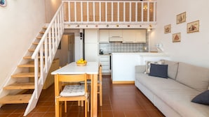 Un espace de vie compact comprenant un escalier menant à un niveau supérieur, un coin repas avec une table et des chaises, une cuisine avec des meubles blancs et un canapé avec des coussins.