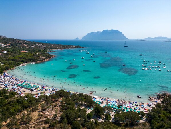 Relax on the beach with umbrellas and sun loungers, surrounded by boats in the turquoise waters and a scenic mountain backdrop.