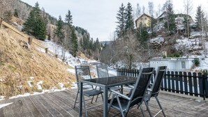 Um deck de madeira com mesa e cadeiras de vidro, cercado por uma cerca de madeira e chão coberto de neve.