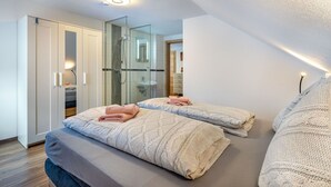 Quarto moderno com duas camas, box de chuveiro em vidro e guarda-roupa branco.