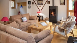 Ein gemütliches Wohnzimmer mit einer Couch, einem Sessel und einem Tisch mit Gläsern und einem Buch. Im Hintergrund ist ein Kamin mit Schornstein zu sehen.