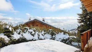 Eine Holzhütte mit schneebedecktem Dach, umgeben von schneebedeckten Bäumen und Bergen im Hintergrund.
