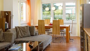 Una sala de estar moderna con un sofá, una mesa de comedor y sillas. Se observa una estantería, un televisor y una ventana con cortinas.