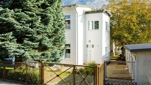 Un edificio moderno de dos pisos con fachada blanca, una entrada porticada y un gran árbol de hoja perenne al frente.