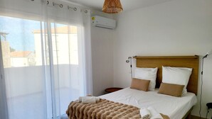 Un letto rifatto con un copriletto e cuscini marroni, una finestra con tende leggere e un condizionatore d'aria a parete.