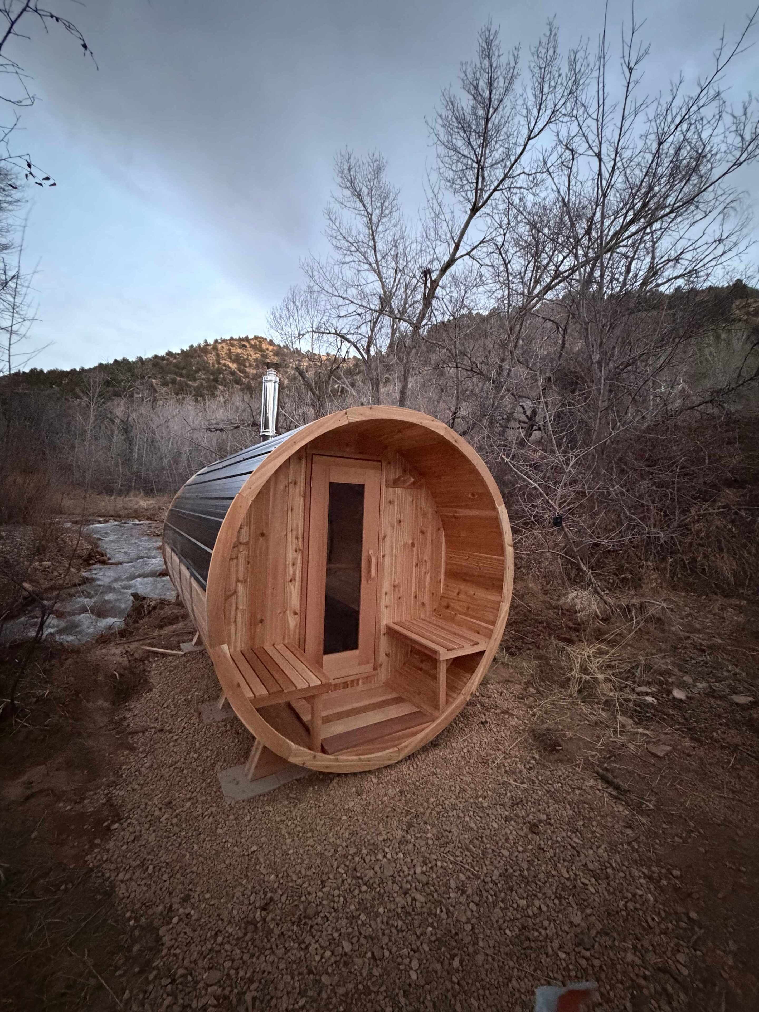 A wooden sauna structure with a door and steps, set in a natural outdoor environment with a river and trees.