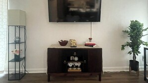 Una sala de estar con un televisor de pantalla plana montado en la pared, una consola de madera oscura con objetos decorativos, una lámpara de pie alta y una planta en maceta.