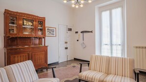 A living room with two striped armchairs, a wooden cabinet with glass doors, a chandelier, a fish decoration on the wall, and a large window with sheer curtains. - Lovely apartment for 4 people with WIFI, TV and balcony (Sestri Levante)