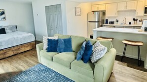 Un espace de vie compact comprenant un lit, un canapé vert avec des coussins bleus, un coin cuisine avec des meubles blancs et un petit tabouret de bar.