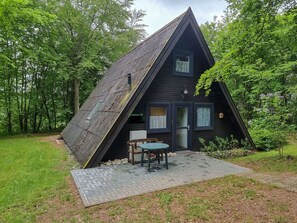A small, dark-colored cabin with a sloped roof, a small porch, and a table and chair set outside.