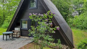 Een zwart a-dak huisje met een klein terras en één raam.