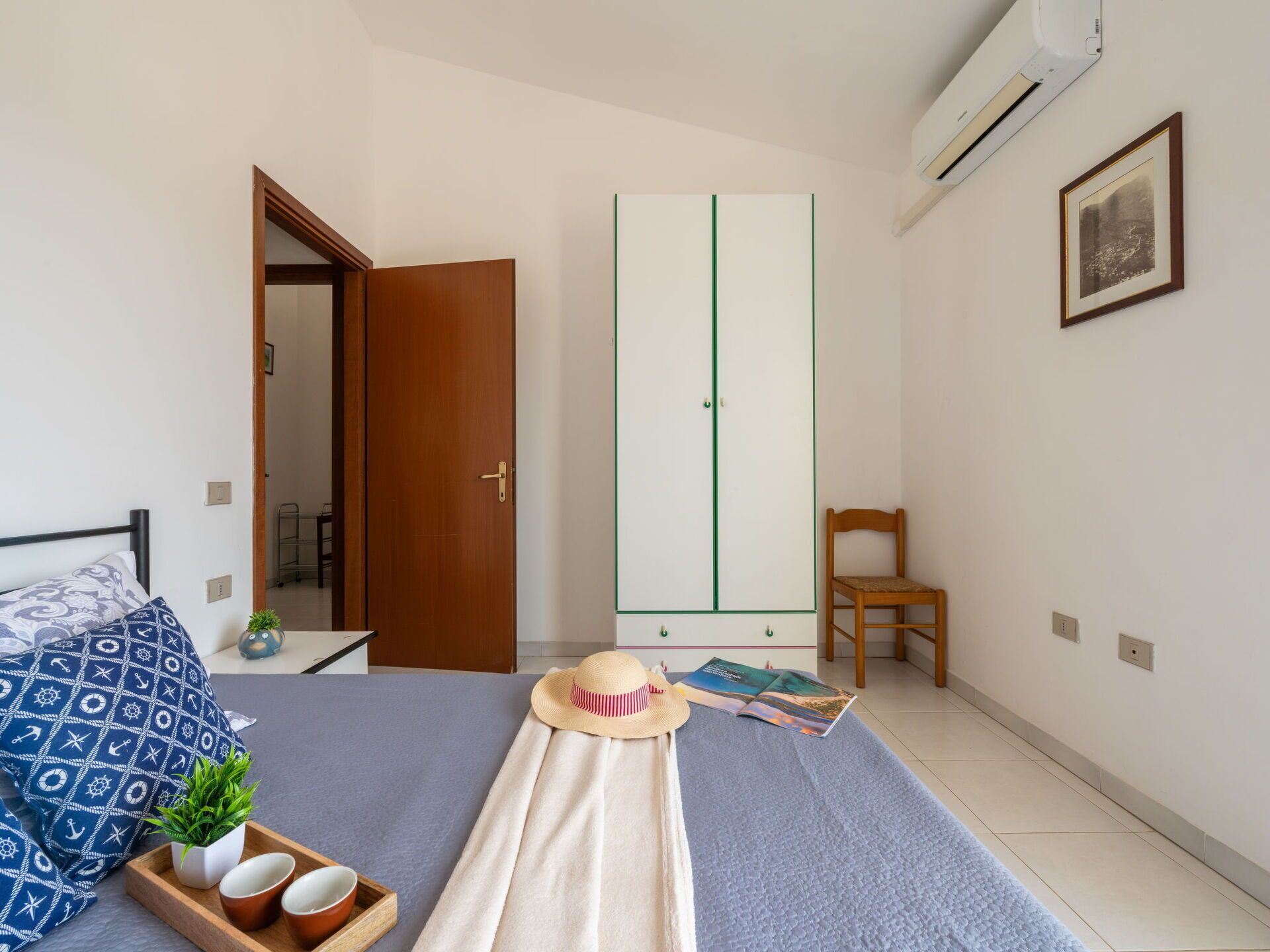 A bedroom with a bed, a wardrobe, a chair, and a small table with a plant and cups.