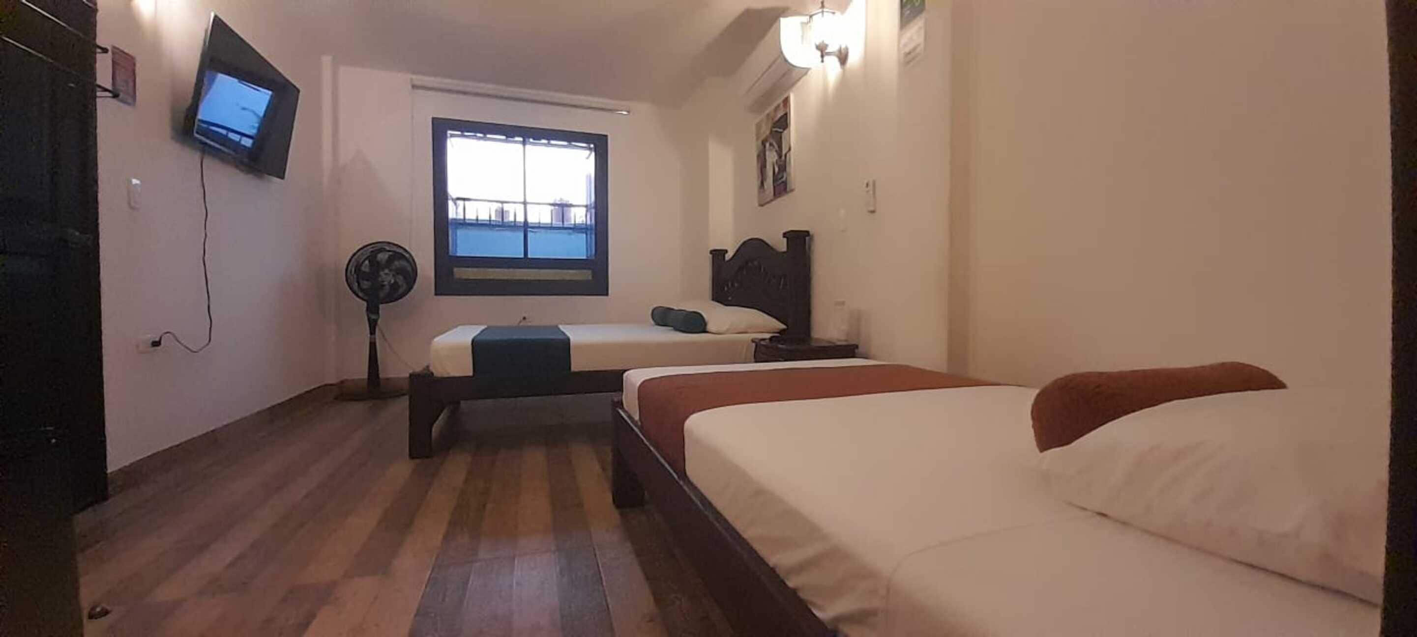 Una habitación de hotel con cama, televisor montado en la pared, ventana y mesita de noche.
