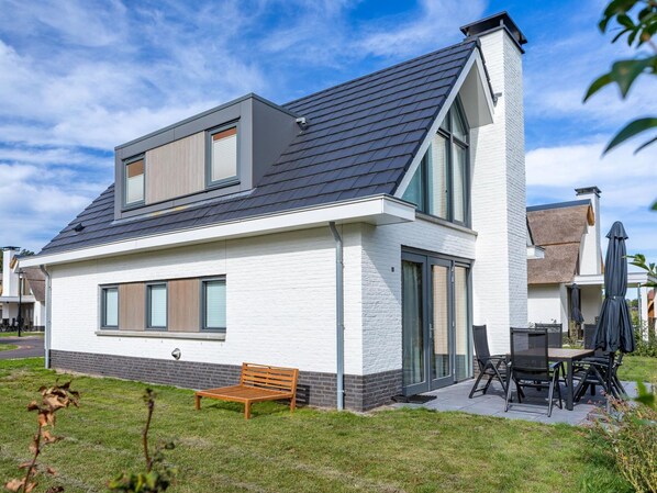 A modern two-story house with a sloped roof, large windows, and a patio area with outdoor furniture.