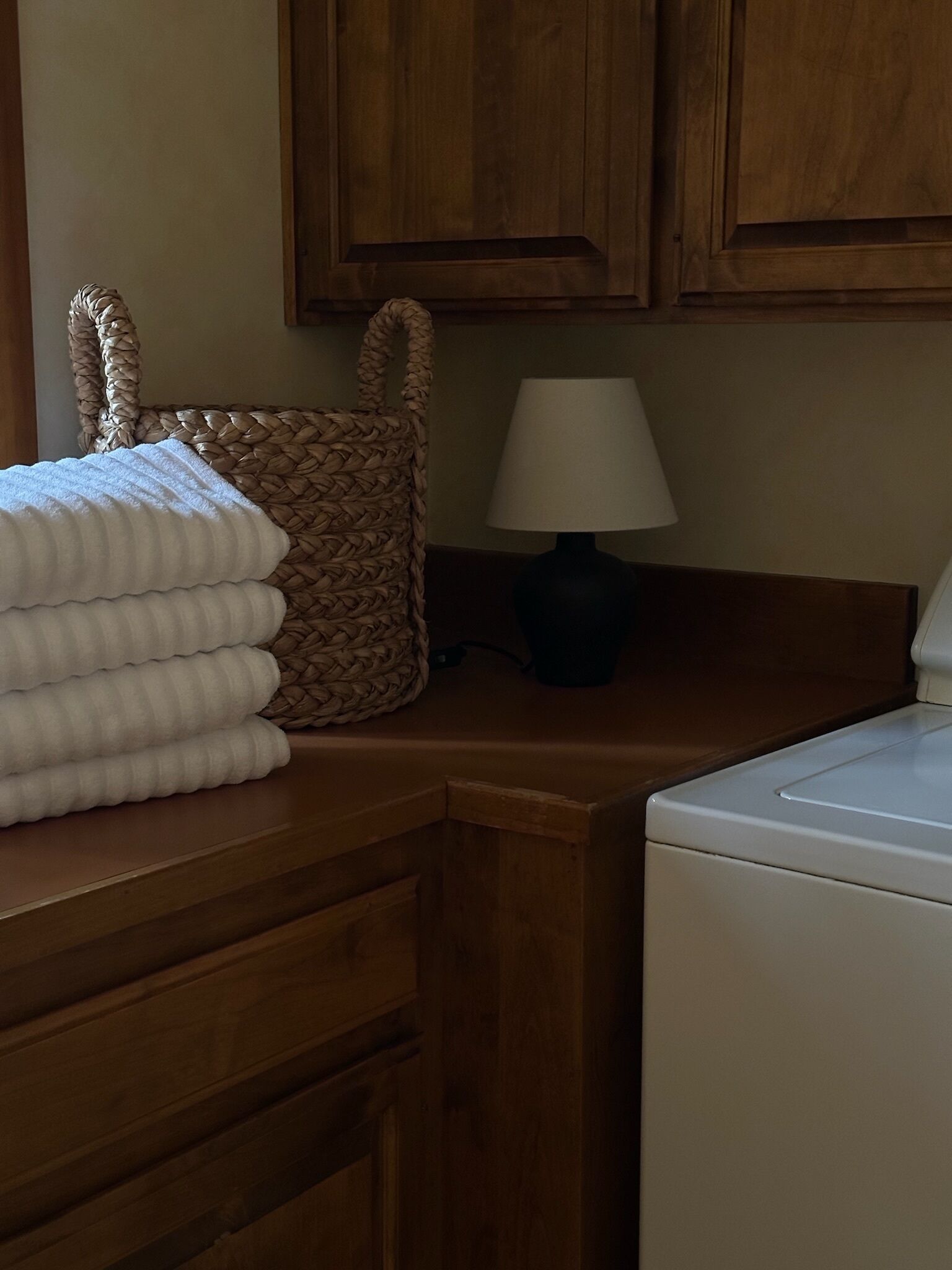 A wooden counter with a woven basket, a stack of white towels, a lamp, and a washing machine.