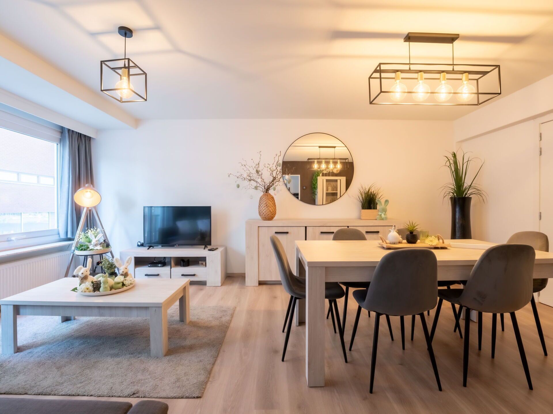 Ein modernes Wohnzimmer mit Fernseher, Esstisch und Couchtisch. Ein großes Fenster mit Vorhängen, eine Pflanze und ein Spiegel an der Wand.