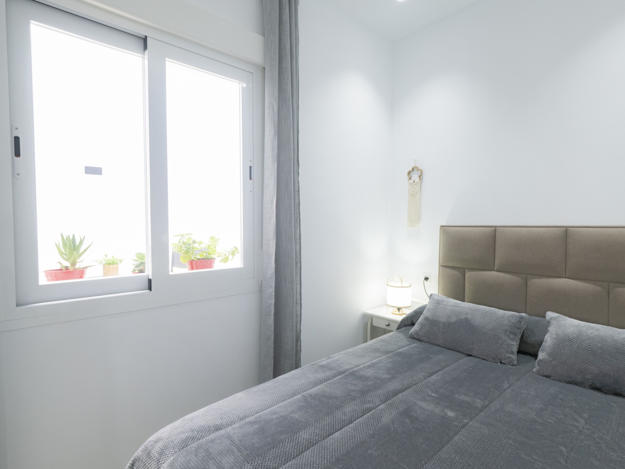 Une chambre à coucher avec un lit, une table de chevet, une lampe et une fenêtre agrémentée de plantes en pot.