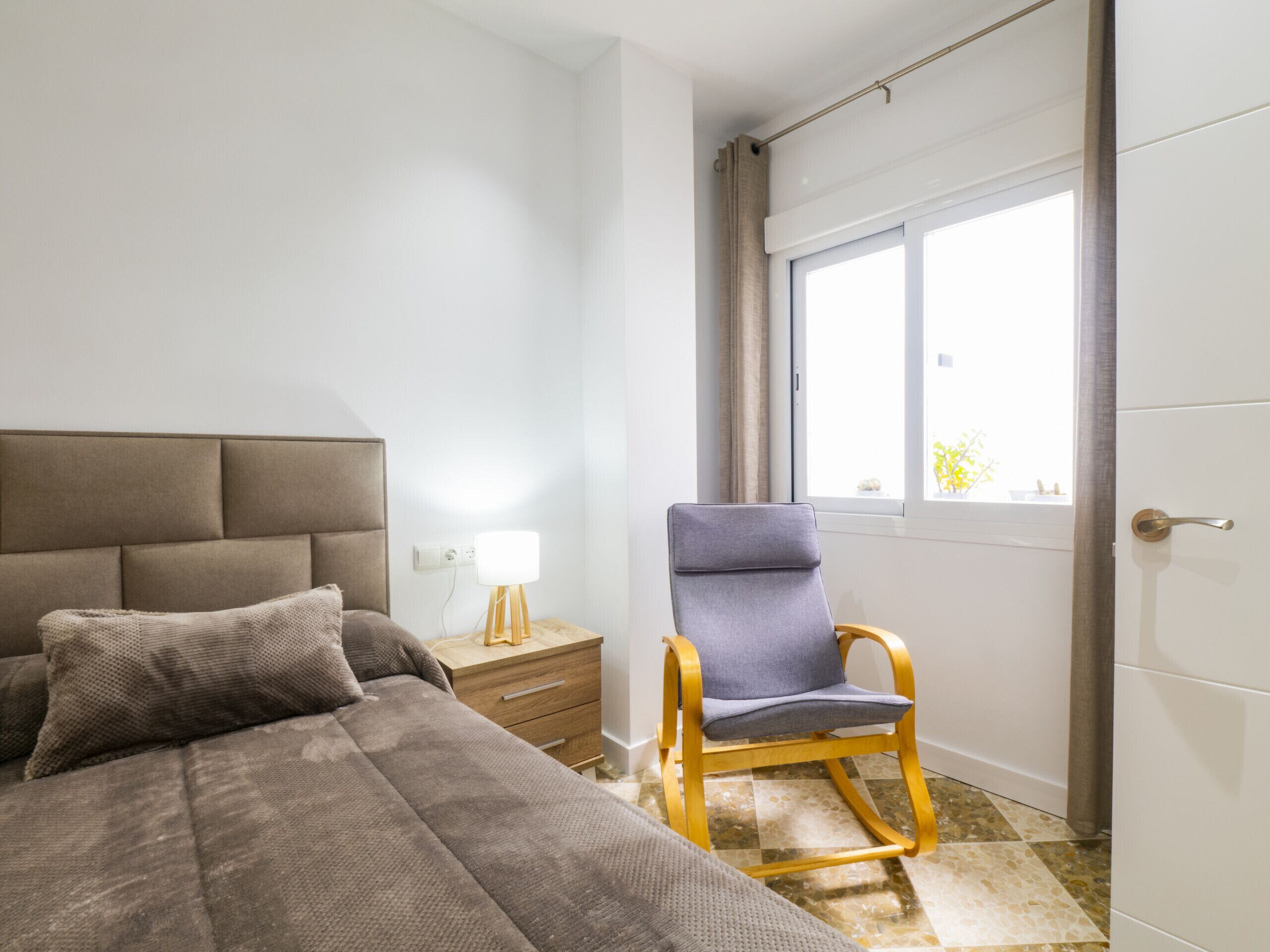 Une chambre à coucher avec un lit, une table de chevet, une lampe, une chaise et une fenêtre.