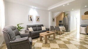 Una sala moderna con un sofá, un sillón y una mesa de centro, un comedor con una mesa y sillas, y una cocina equipada.