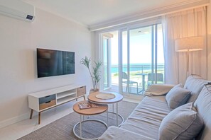 A modern living room with a flat-screen TV, a sofa, two small tables, and a large sliding glass door leading to a balcony with a view of the sea. - Oceana Suites Cruceros III (Punta del Este)