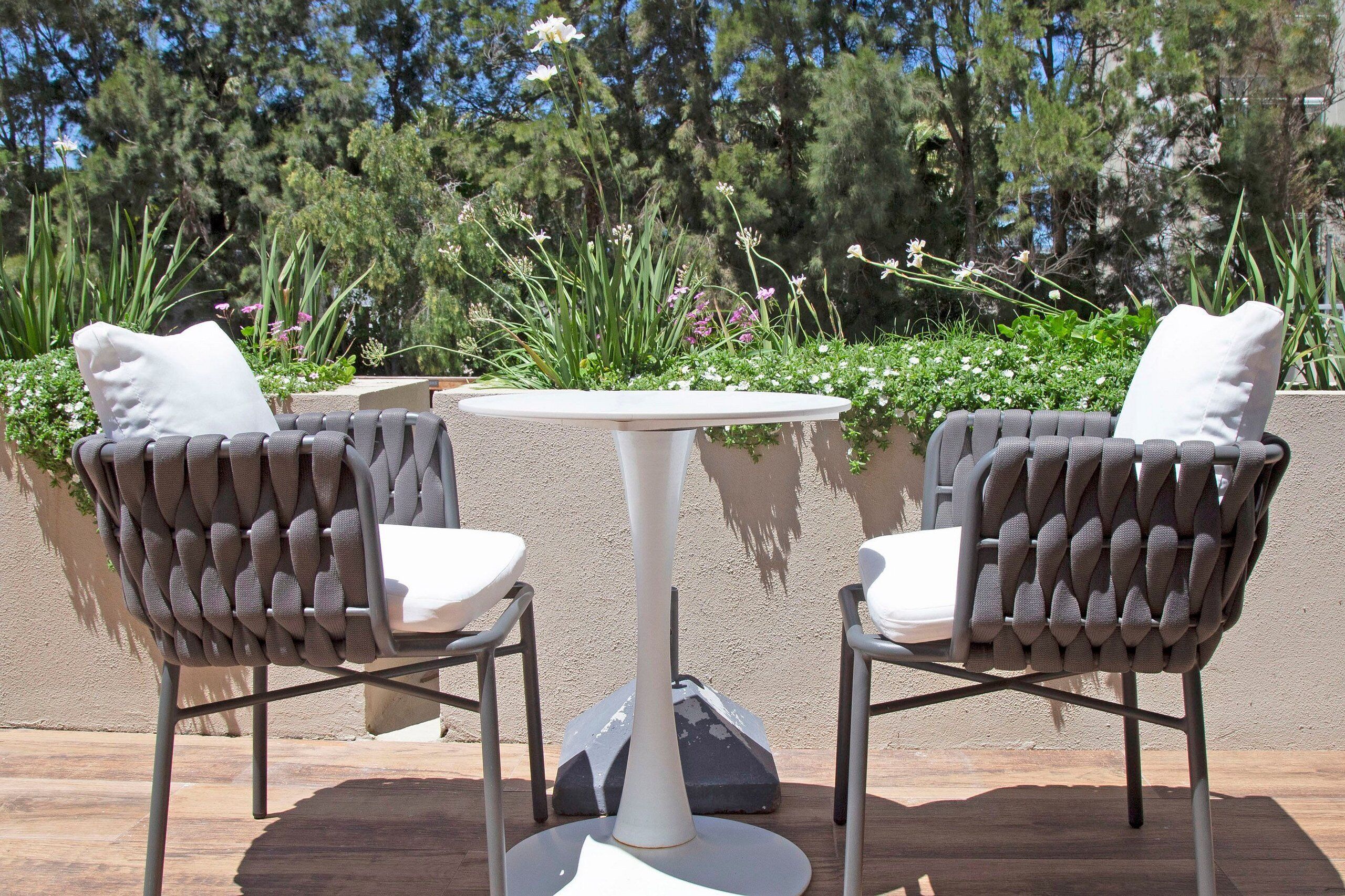 Two chairs with white cushions and a white table, set outdoors with a view of greenery and trees.
