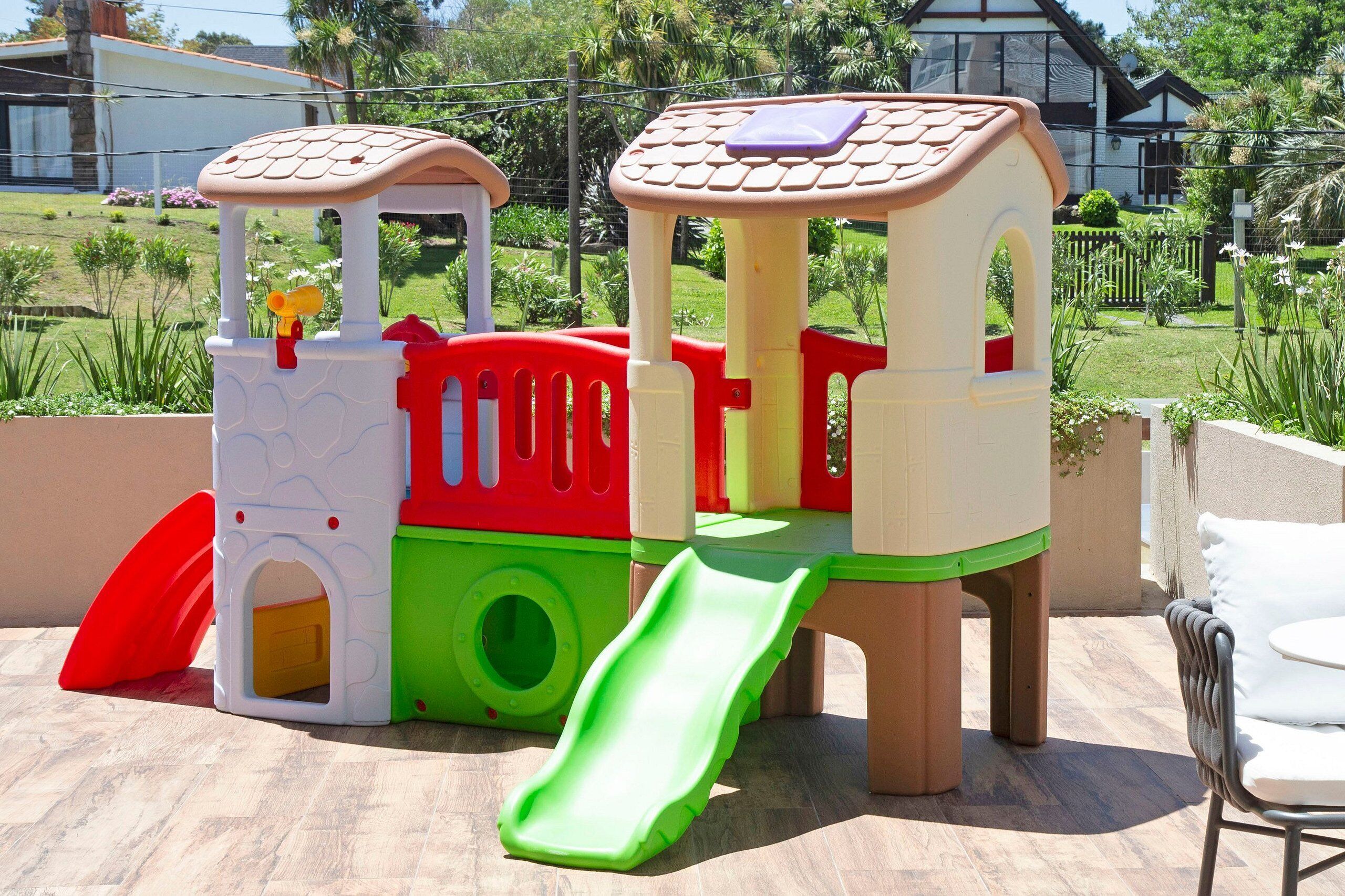 Une aire de jeux pour enfants, avec des toboggans et des zones d’escalade, installée dans un jardin, avec une maison et un autre jardin en arrière-plan.