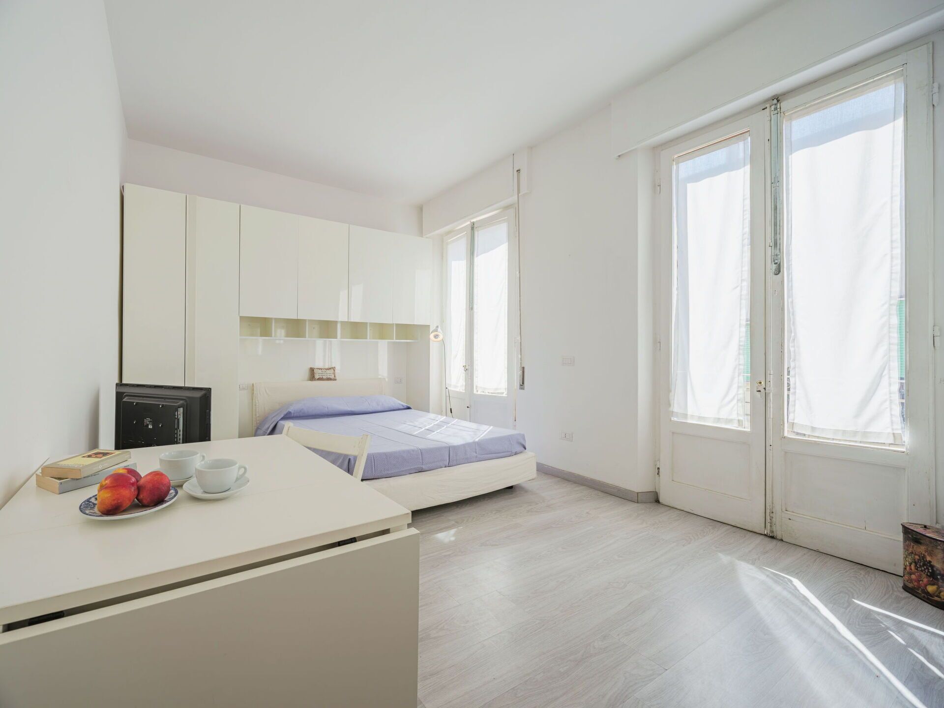 Een moderne slaapkamer met een bed, een bureau met fruit en grote ramen.
