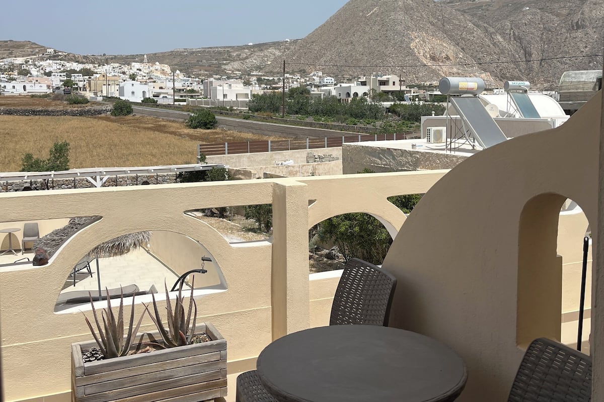 A balcony with a table and chairs, overlooking a town with white buildings and a mountain.