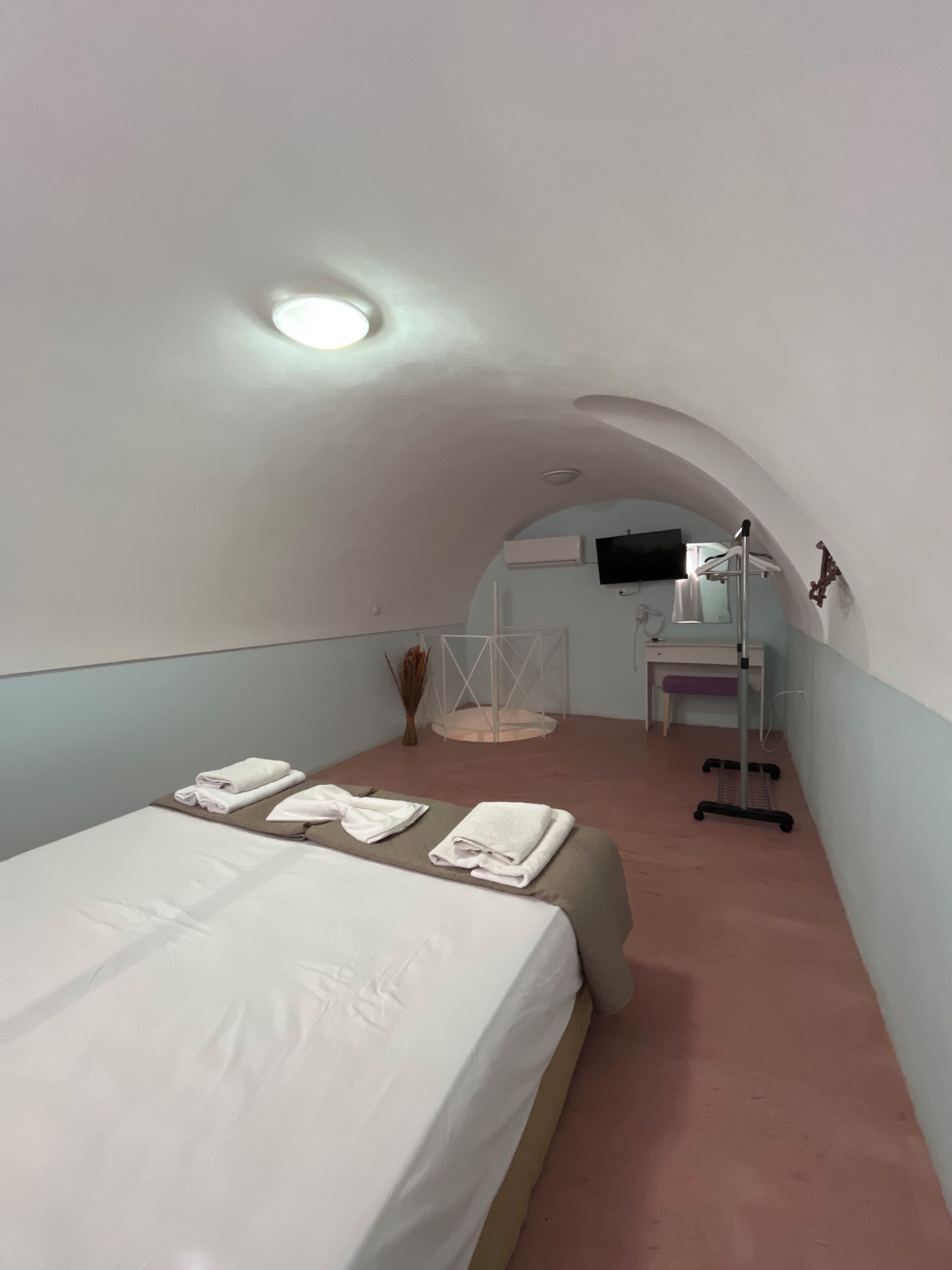 A small, cozy room with a single bed, a desk, and a chair. There is a mirror on the wall and a small table with a lamp.