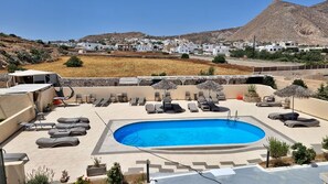 A pool area with lounge chairs and umbrellas, surrounded by a stone wall and a view of a hillside town. - Leon Lux Apartments (Santorini)