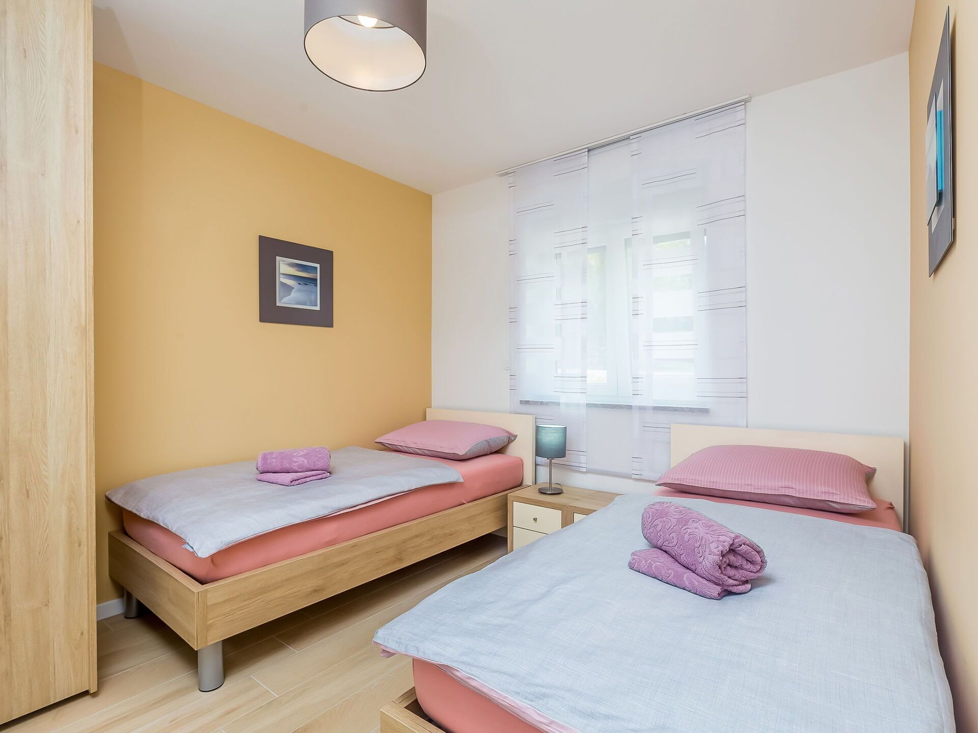 A bedroom with two beds, a bedside table, a lamp, and a window with blinds.