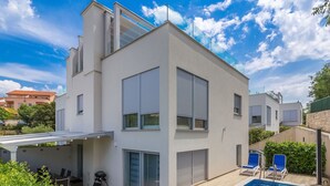 Ein modernes, zweistöckiges Haus mit Pool, überdachter Terrasse und Blick auf Nachbargebäude.