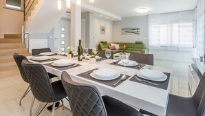 Ein moderner Essbereich mit einem weißen Tisch, gedeckt für vier Personen, schwarzen Stühlen und hängenden Pendelleuchten. Im Hintergrund ist eine Treppe zu einer oberen Etage zu sehen.