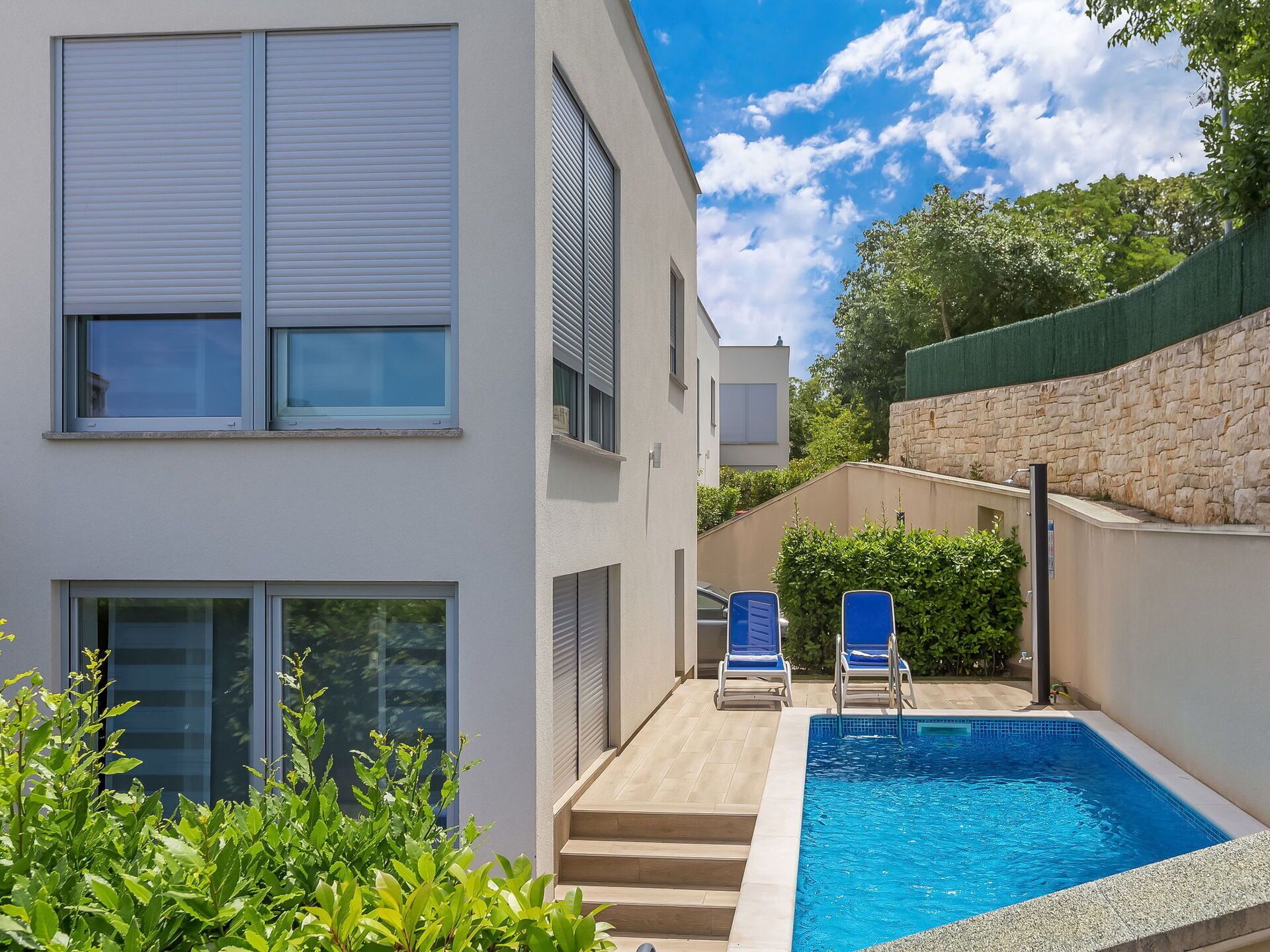 A modern building with a swimming pool, two lounge chairs, and a stone wall.