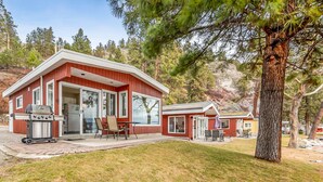 A two-story cabin with a red exterior, large sliding glass doors, a patio with outdoor furniture, and a stone wall.