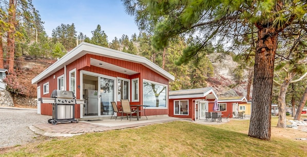 A two-story cabin with a red exterior, large sliding glass doors, a patio with outdoor furniture, and a stone wall. - Perfect Getaway Cabin in the Okanagan 6 (West Kelowna)