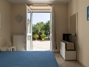 Ein Schlafzimmer mit Bett, Fernseher, Kommode und einem Fenster zum Balkon mit Topfpflanzen.