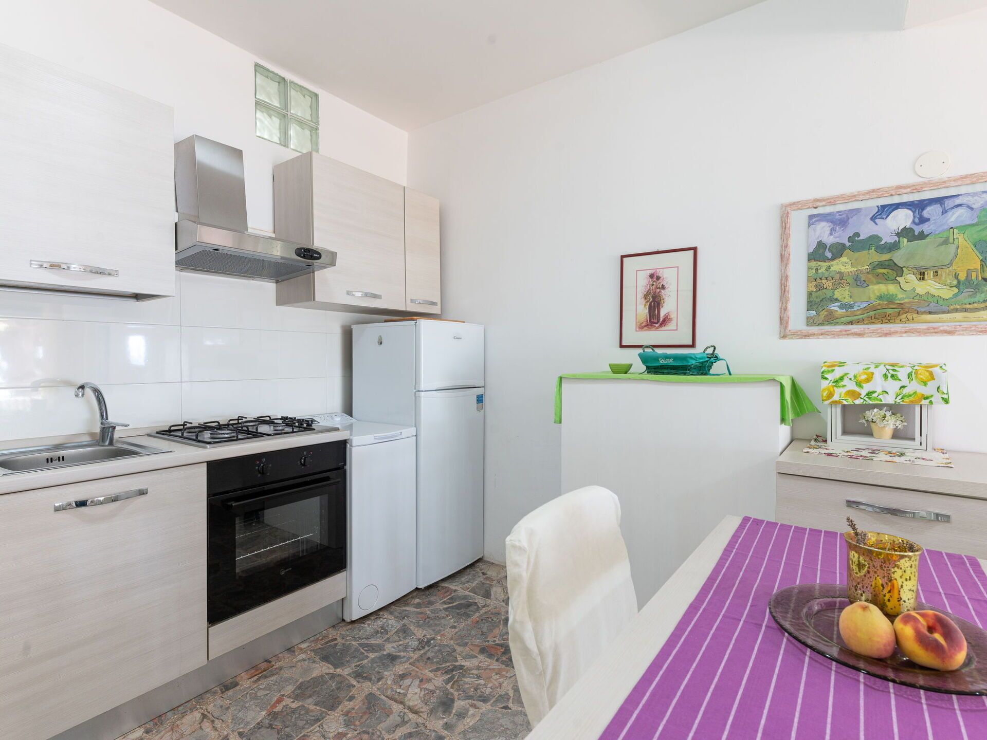 Une cuisine moderne avec des meubles blancs, une cuisinière à gaz, un four et un réfrigérateur blanc. On y trouve une table à manger recouverte d’une nappe violette et une assiette de fruits.