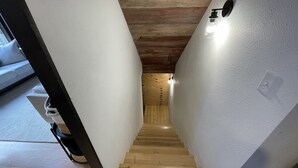 Un couloir étroit, doté d’un plancher en bois et de murs blancs, mène à une pièce où l’on aperçoit un interrupteur et un luminaire au plafond.