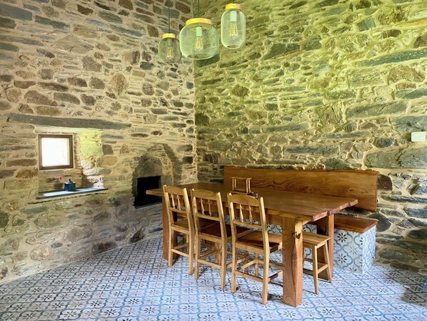A rustic stone-walled room with a wooden table and chairs, a small window, and a stone fireplace.